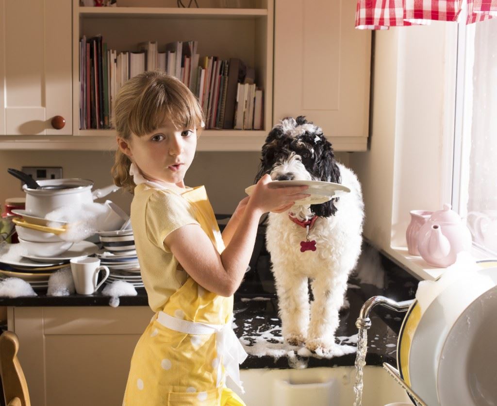Little girl holding plate for dog to lick while doing the dishes